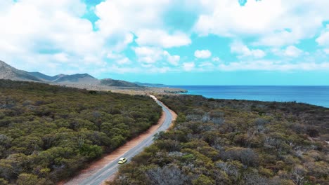 El-Dron-Sigue-Un-Automóvil-Amarillo-Que-Conduce-Por-La-Naturaleza-En-Una-Pequeña-Carretera-Cerca-Del-Océano-En-Imágenes-Aéreas-De-Curacao-4k