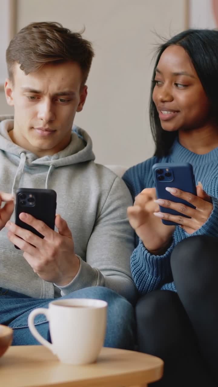 Vertical video: Starting texting, couple in hoodie and blue knit tapping screens at home with mug