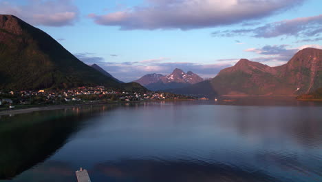 aerial panning right shot of a small village on the slopes of a hill along a fjord in northern norway with mountains and clouds in the background at sunset