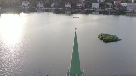 Cruz-Encima-Del-Campanario-De-La-Iglesia-Libre,-La-Iglesia-Libre-En-Reykjavík,-Agua-Del-Lago-Tjörnin