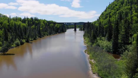 Un-Dron-Vuela-Sobre-El-Agua-De-Un-Río-Con-árboles-Y-Montañas-A-Ambos-Lados-Y-Un-Viejo-Puente-Ferroviario-Al-Fondo