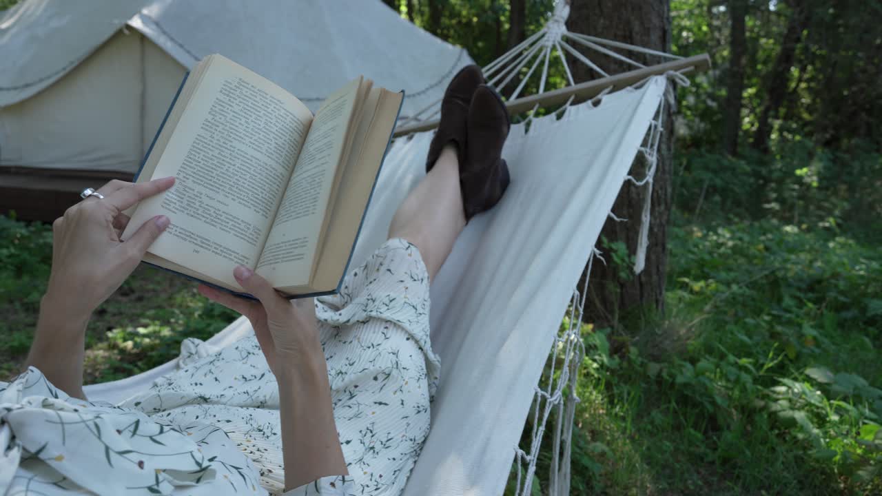 Girl with dark hair in white country dress reads a book in a hammock (view 3)