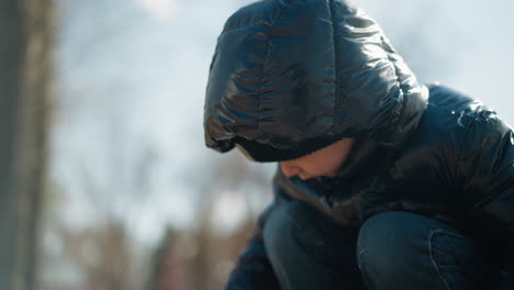 a young boy deeply focused on his activity, wearing a shiny black jacket, a head warmer, and jeans, with a blur background of trees