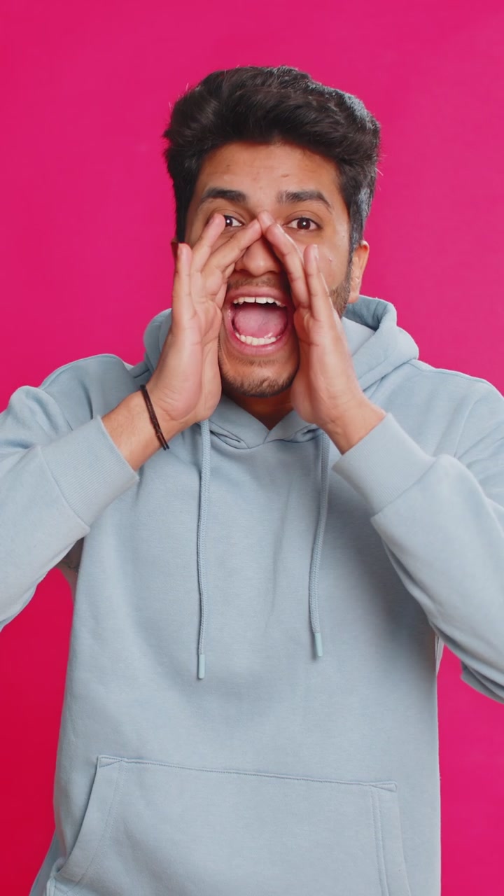 Indian young man shouting calling out loud holding hands near mouth yelling waving excited indoors