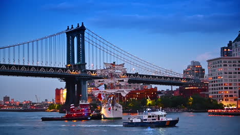 ship with white sails, tugboat and police boat on the river. the manhattan bridge with lights on at dusk time at backdrop