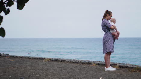 contra el panorama del mar y la playa, un hermoso retrato captura el suave beso de una madre otorgado a su hija. la familia de vacaciones usa expresiones de felicidad, y la madre sostiene a su bebé cerca