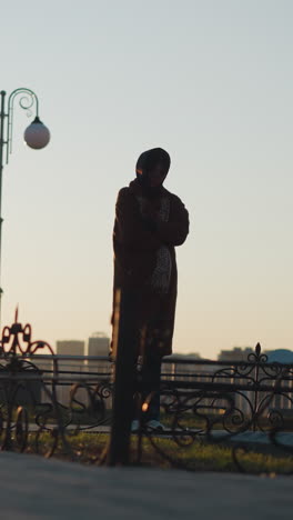a mournful woman wearing a coat and scarf stands in front of a stone monument in a park during sunset. sorrowful stance, with the warm, fading light of the setting sun casting long shadows