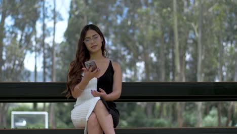 Attractive-young-girl-in-a-dress-is-sitting-and-smiling-at-the-camera-when-her-phone-rings-and-she-starts-talking-cheerfully