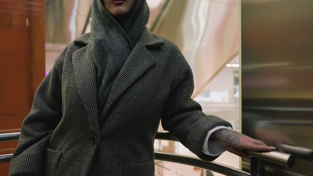 woman in checkered coat and hooded scarf rides glass elevator in modern mall, ascending slowly with calm expression as urban lights and reflections pass behind transparent panels in shopping center