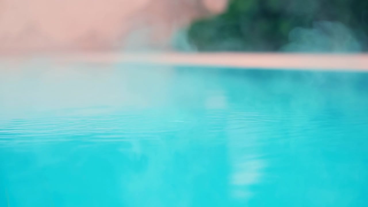 Pemandangan sudut rendah air kolam pirus bening dengan riak lembut, kilau halus, dan cahaya siang yang lembut, menunjukkan gerakan tenang di seluruh permukaan bersih di samping bibir kolam berwarna persik pucat dan tanaman hijau yang buram.