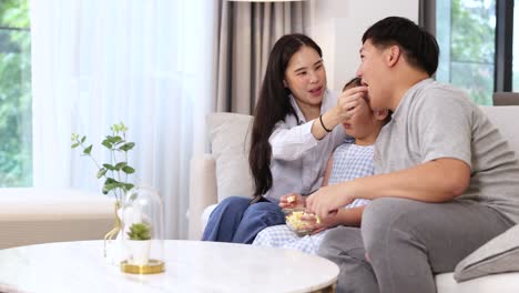 a family of three shares popcorn on a cozy sofa, creating a warm and joyful atmosphere in a bright living room