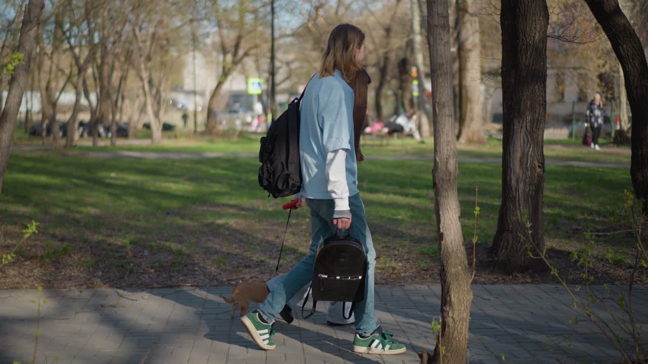 joven adulto paseando a un perro en un parque, persona que se desplaza en monopatín lleva una tabla y una bolsa mientras un perro pequeño explora la hierba, parque urbano de fondo con bancos y gente a la distancia, ambiente relajado de transporte