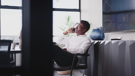 young black man talking on phone in an office, feet up on table, close up, seen through glass wall