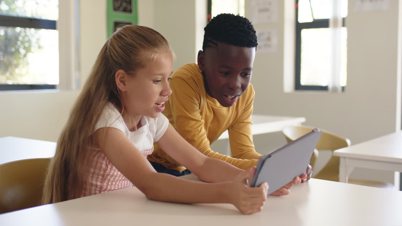Diverse youth classmates in classroom, girl angling tablet, boy in mustard top leaning in, smiling