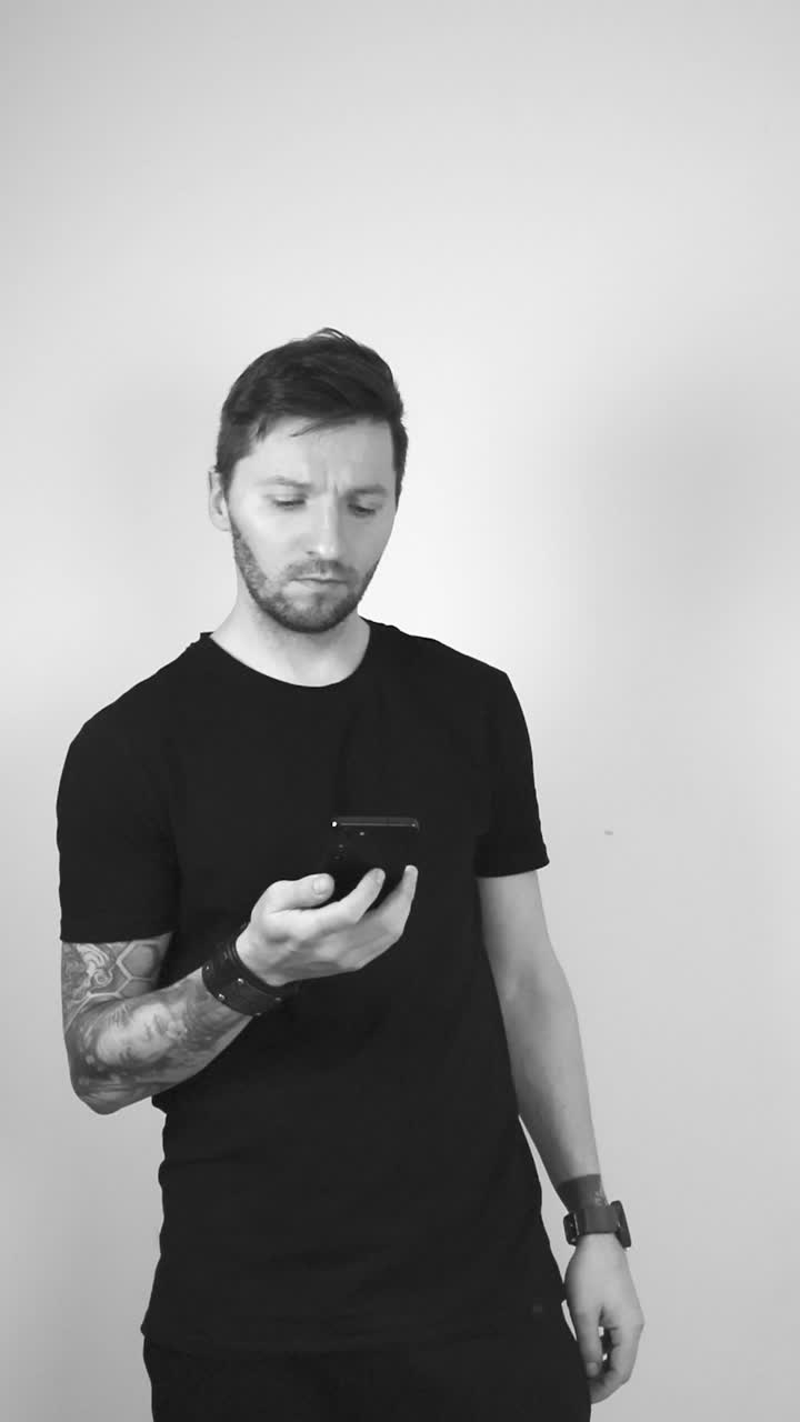 Grayscale footage of a white tattooed man with short hair and beard scrolling through his phone or smartphone in slow motion showing that he is in social media or browsing something. Concerned face.