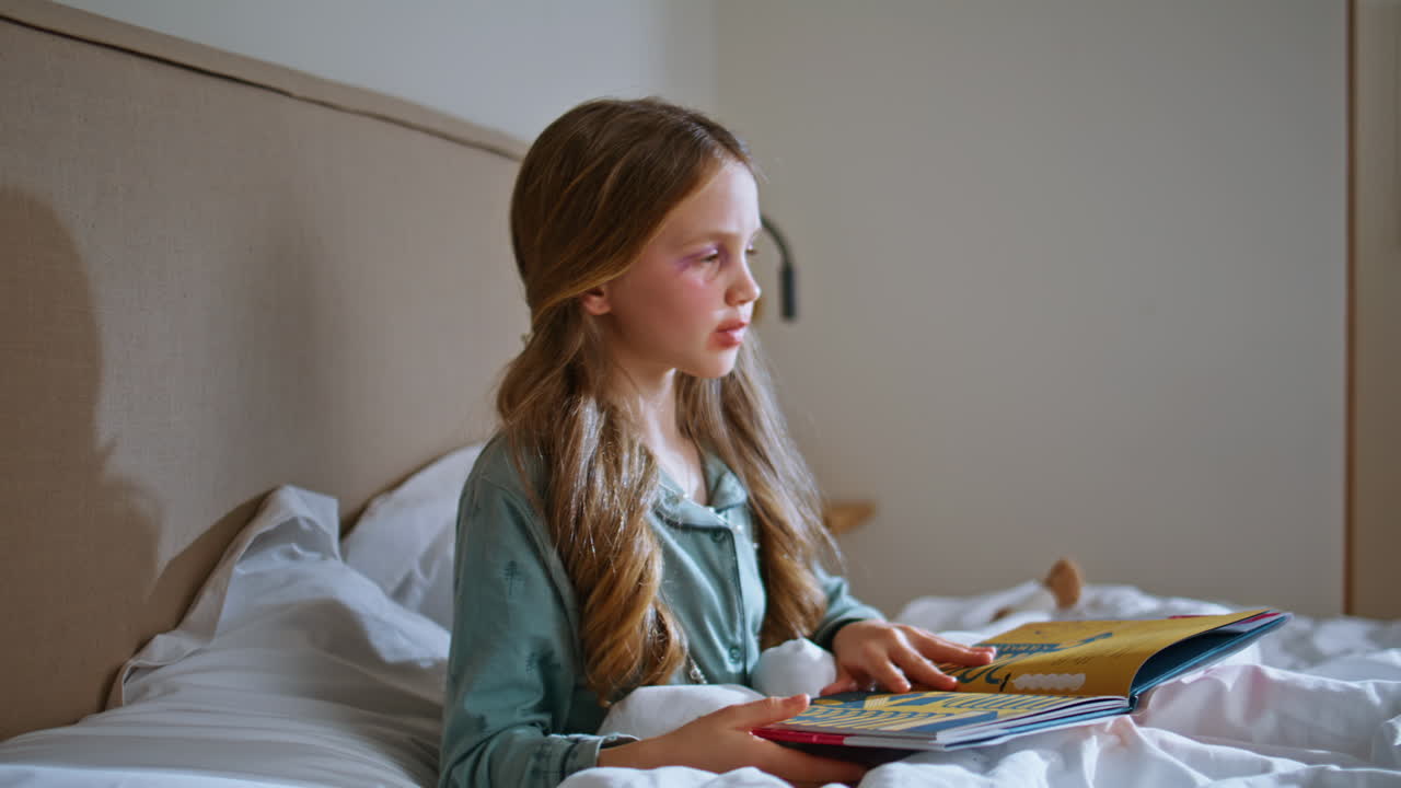 自宅の柔らかいベッドに座って本を読んでいる、のんきな赤ちゃんのクローズアップ。子供がページをめくっています。