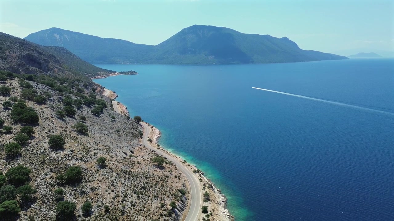 Sahil yolu üzerinde alçalan sinematik drone görüntüsü, arka planda büyük dağlarla sakin mavi denizde seyreden bir sürat teknesini ortaya çıkarıyor