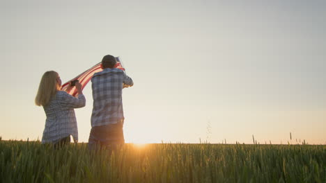 Wir-Feiern-Den-Unabhängigkeitstag-–-Ein-Mann-Schwenkt-Die-US-Flagge,-Eine-Frau-In-Der-Nähe-Fotografiert-Mit-Einem-Smartphone.-Stehend-Vor-Dem-Hintergrund-Eines-Weizenfeldes,-Wo-Die-Sonne-Untergeht