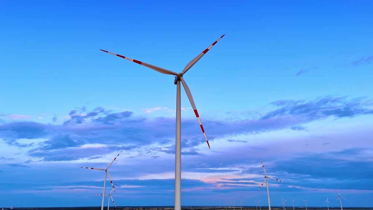 Turbina eolica ravvicinata che domina il cielo limpido e blu del crepuscolo. La vista dal basso mostra pale contrassegnate di rosso e numerosi generatori più piccoli lungo l'orizzonte piatto.