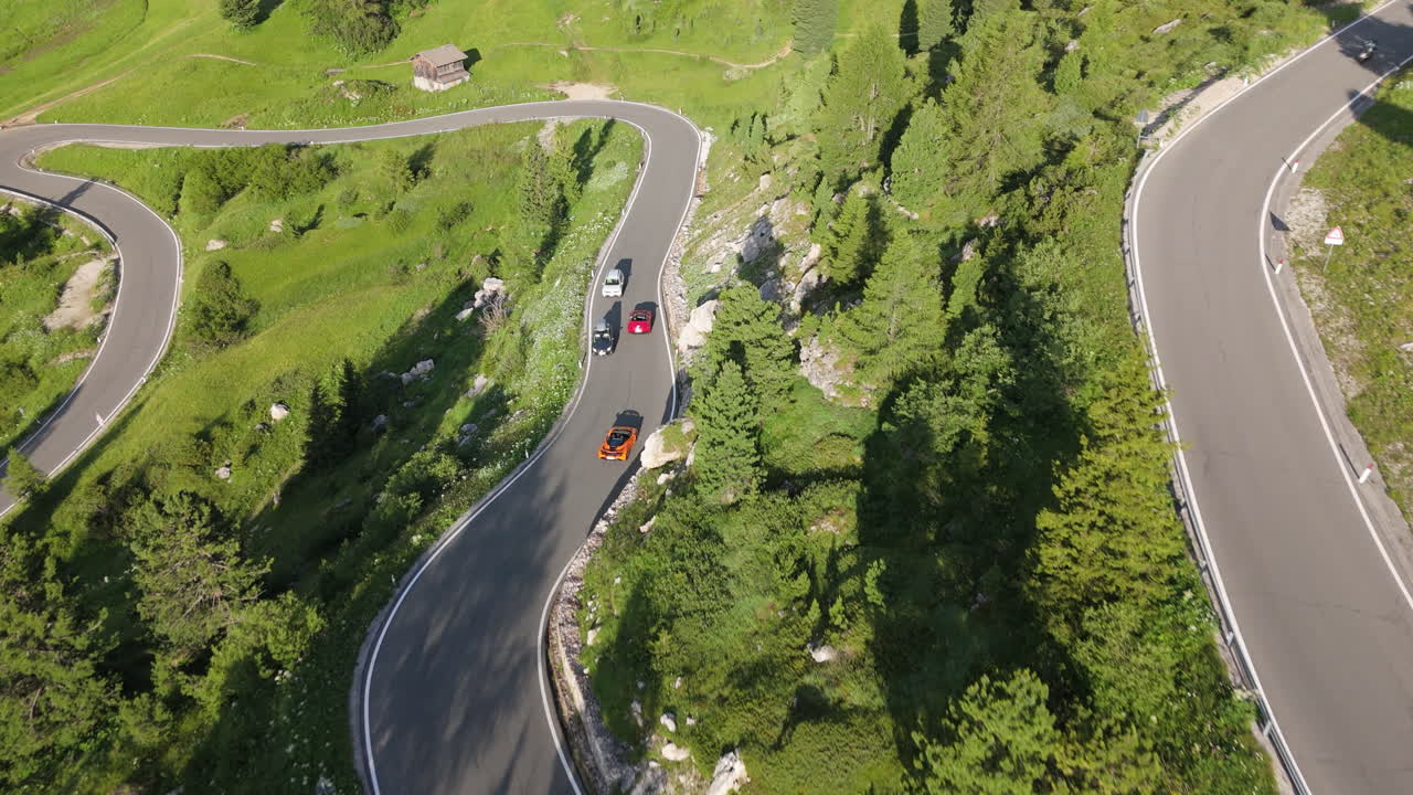 Drone follows three sports cars on a winding alpine road at sunrise, with lush green meadows and forest slopes in view