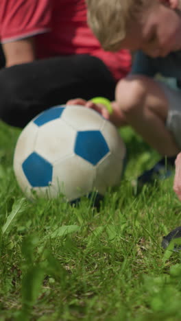 close-up of four people in an outdoor setting, with three kneeling down to tie their shoelaces while a little boy places his hand on a soccer ball