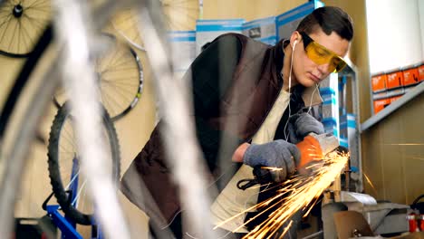 focused repairmen in protective glasses is working with electric circular saw in studio with wooden walls and spare parts on shelves. bicycle parts are in foreground.