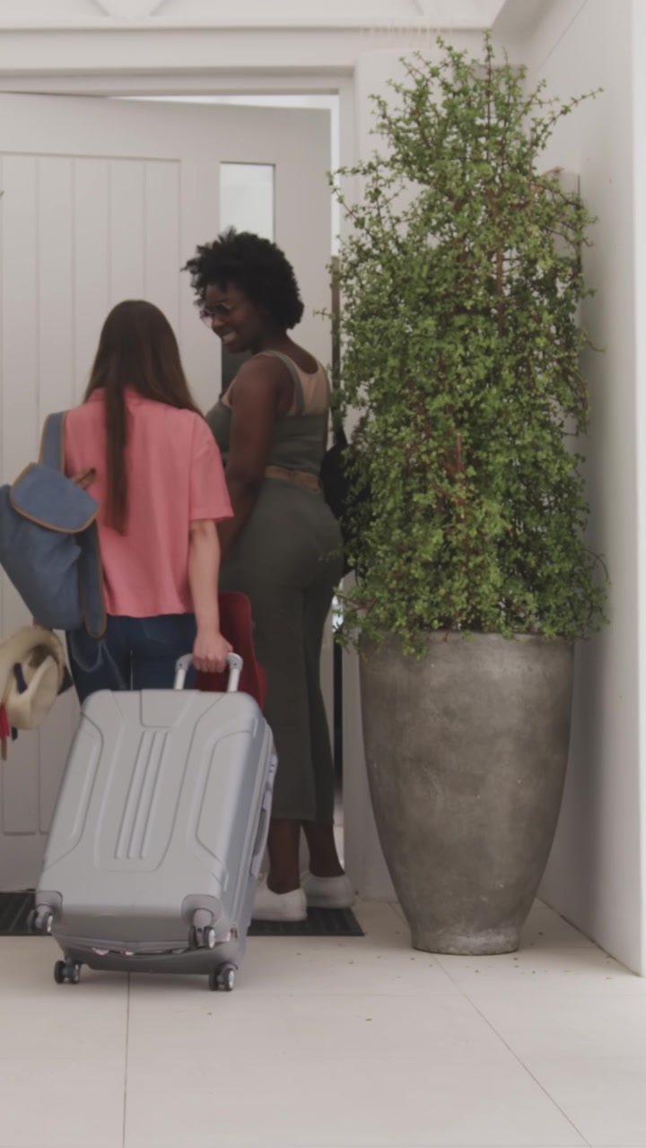 Vertical video: Diverse female friends arriving in foyer, pulling suitcase, pointing, entering home