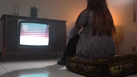 back view of woman sitting on pillow on the floor and watching retro tv with flickering green screen monitor, lonely person being alone at home sitting in front of old television with direct broadcasting. old vintage television been screen, woman watching