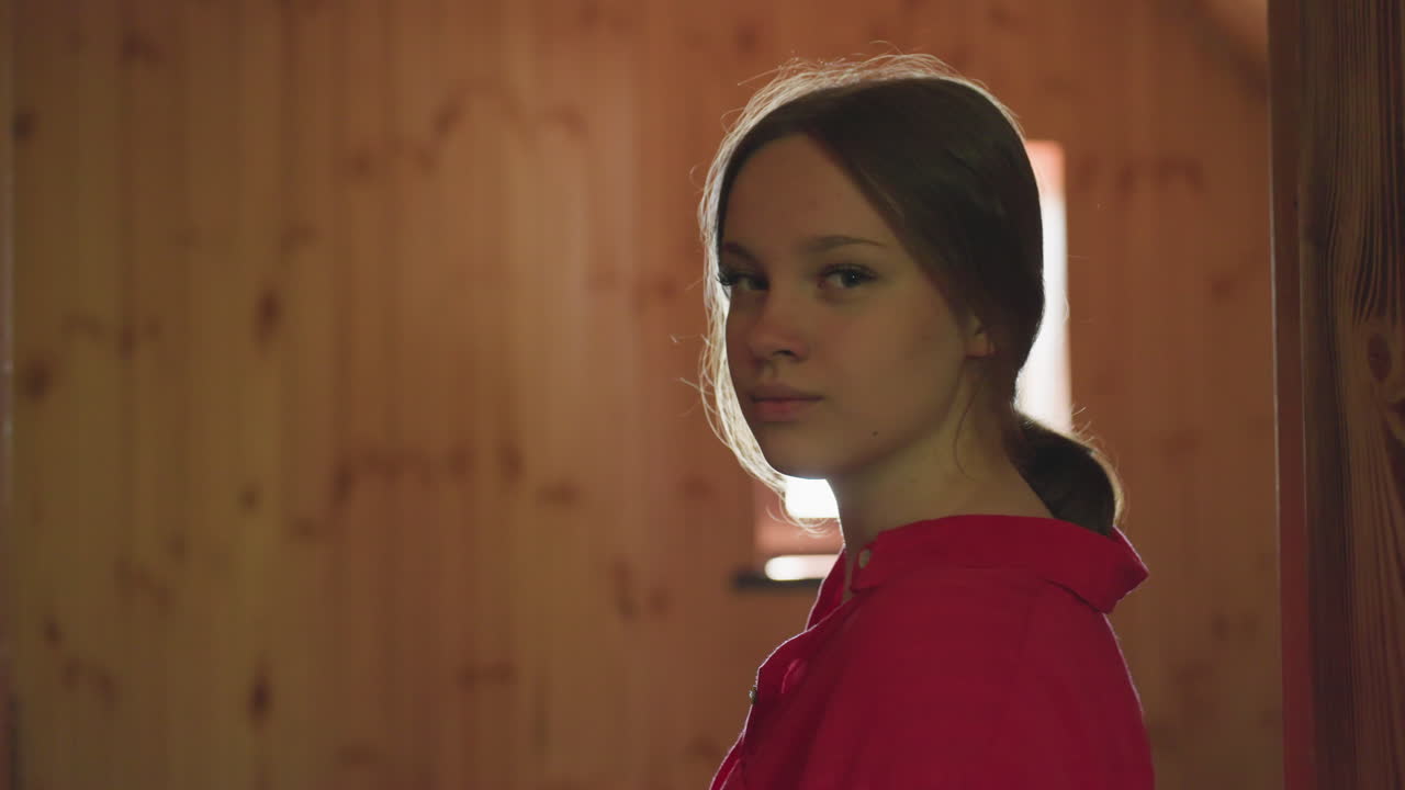 young woman stands quietly near wooden room doorway then turns back gently as warm window light streams in, creating calm and reflective atmosphere in rustic space