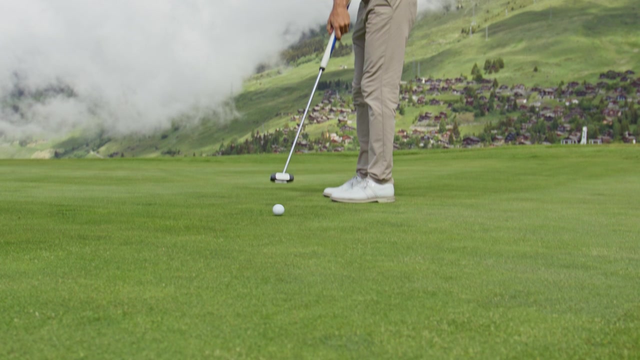 Inquadratura cinematografica dal basso di una pallina da golf che rotola sul green e cade nella buca su un campo da golf di montagna a Verbier, Svizzera. Il paesaggio alpino e un putting di precisione creano un'esperienza sportiva di alta qualità.