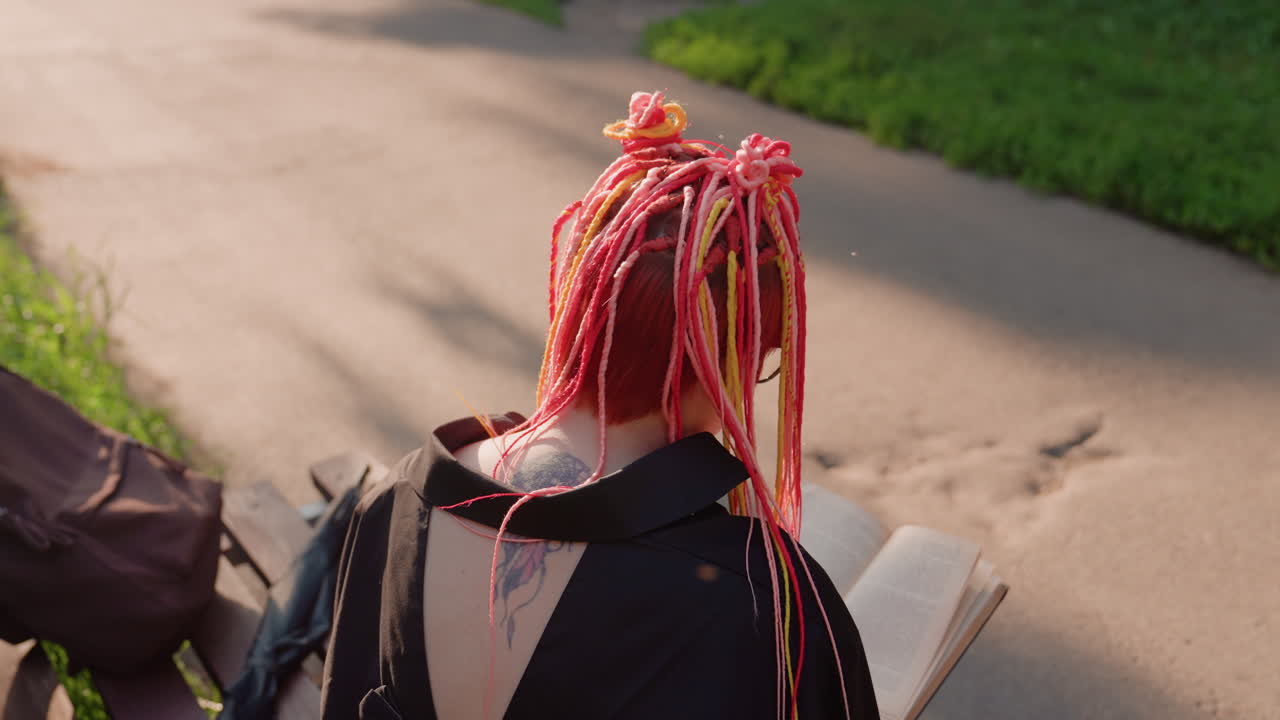 Mujer con trenzas rosas relajándose al aire libre, persona tranquila con pelo colorido absorta en la lectura en el exterior, escena serena de una persona con trenzas vibrantes absorta en la lectura al aire libre en un día soleado