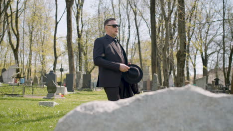 sad man in black suit and sunglasses walking in a graveyard in a sunny morning