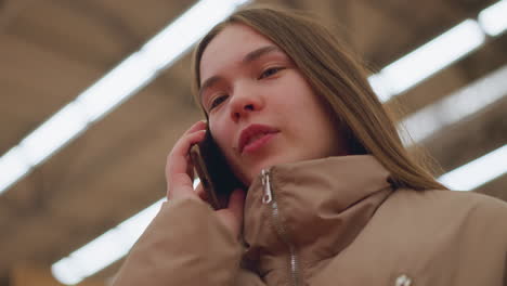 close-up of a girl wearing a brown jacket, standing in a mall, holding a phone to her ear while talking and smiling. the background is blurred, emphasizing her joyful expression of her conversation