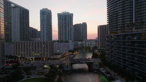 Aerial-ascending-footage-of-high-rise-buildings-in-modern-urban-borough-at-twilight.-Cars-driving-on-bridge-over-river.-Miami,-USA