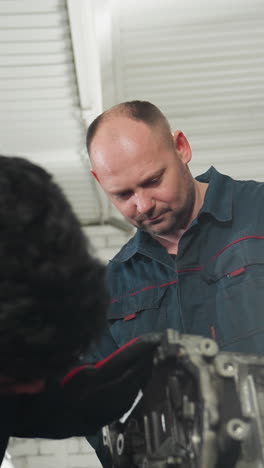 dos ingenieros automotores en un taller, uno intensamente centrado en el ensamblaje de un motor mientras su colega observa atentamente, rodeado de herramientas mecánicas