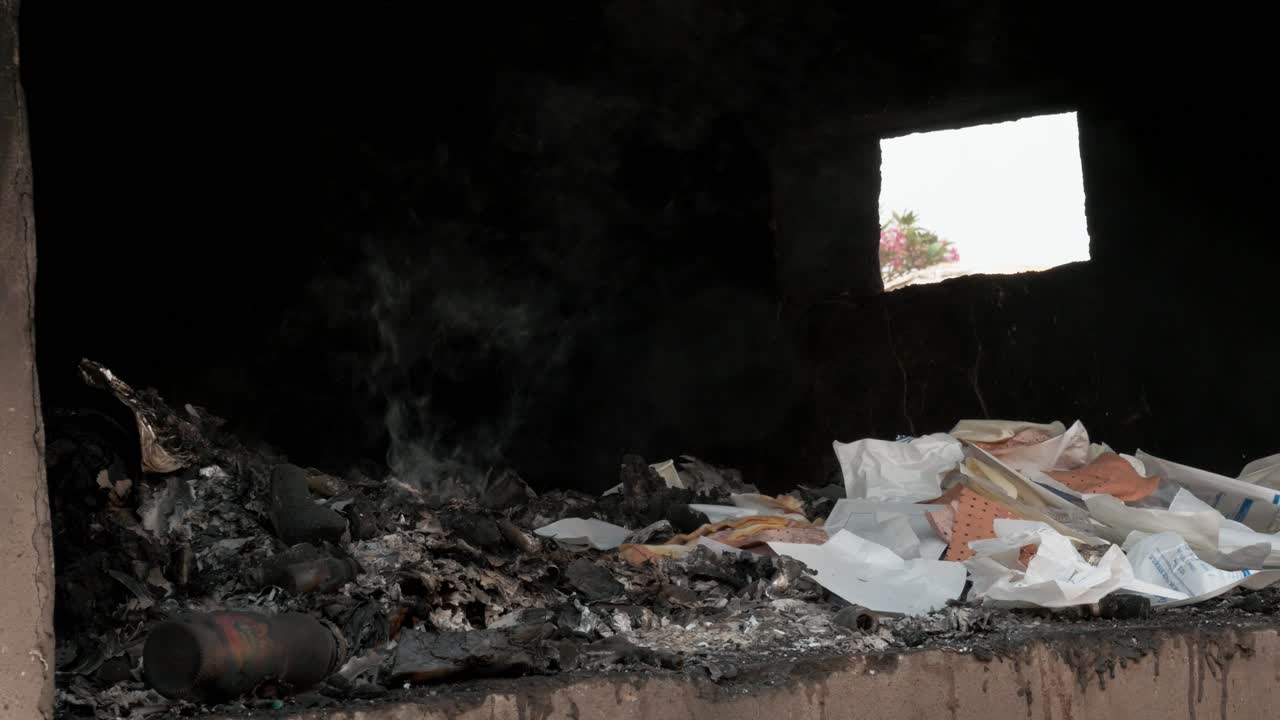 Interno scuro di una struttura piena di rifiuti fumanti e detriti bianchi a Yaoundé, Camerun, che evidenzia i problemi di inquinamento urbano. Inquadratura ampia