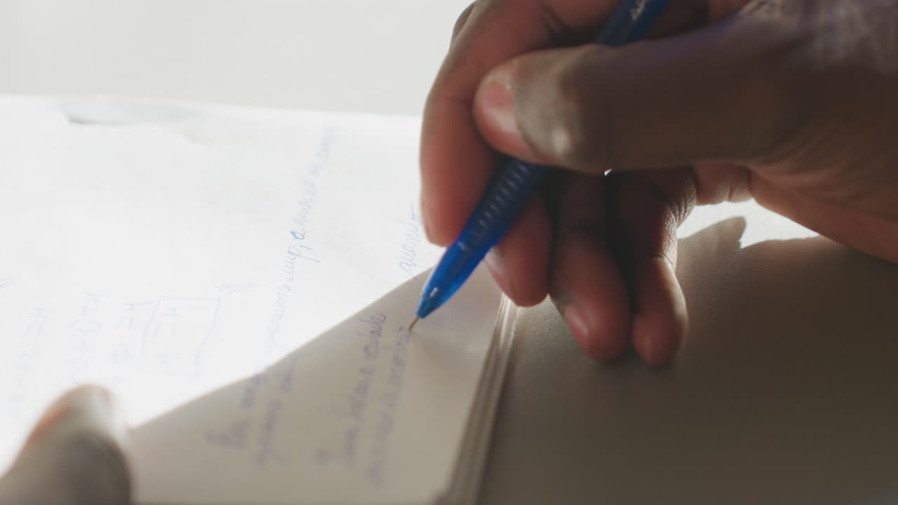 Close up of student hand holding blue pen writing in notebook under soft sunlight, math notes visible on paper, shallow depth of field and shadow across page conveying focus