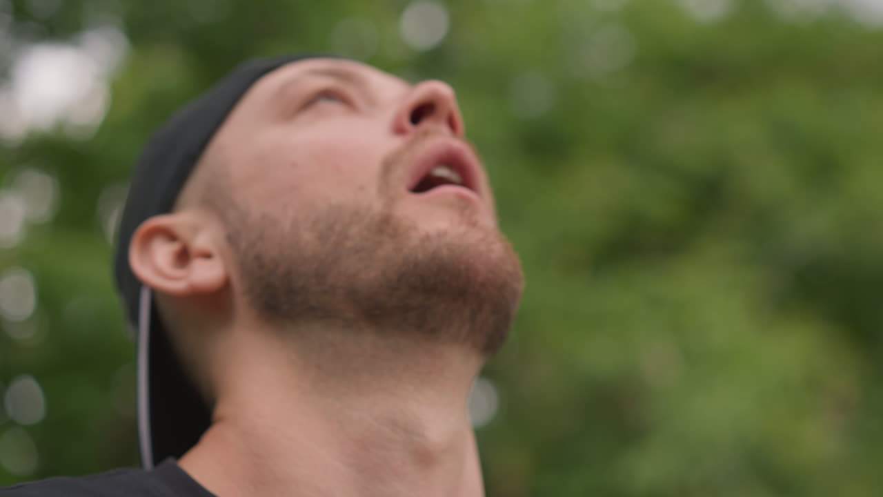 Closeup Breath Caucasian Man Relief After Intense Set, Deep Exhale And Upward Gaze, Cap Backward And Trimmed Beard, Outdoor Green Background, Panting And Pause Between Reps, Candid Portrait Showing