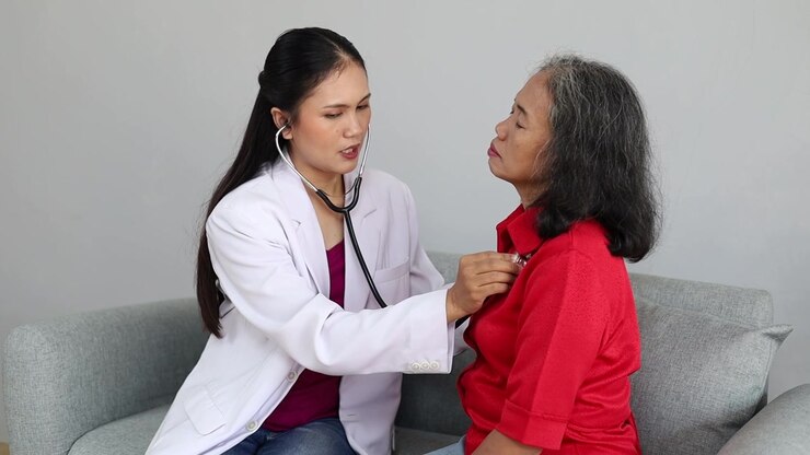 Female doctor in white coat uses a stethoscope to examine an elderly Asian woman at home, representing healthcare, elderly care, medical checkup, and home care.