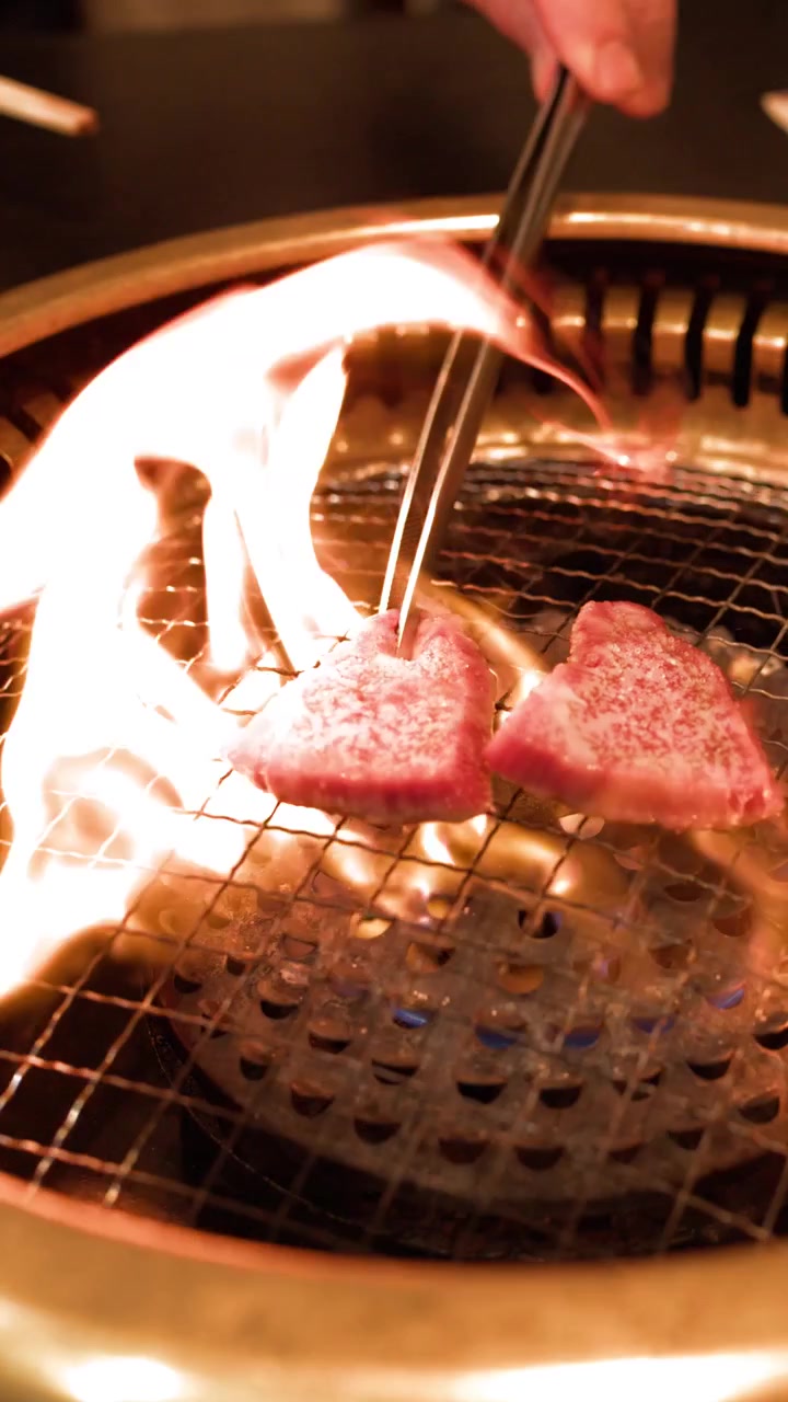 Primo piano di carne di manzo marmorizzata che cuoce su una griglia yakiniku con fiamme intense e pinze.