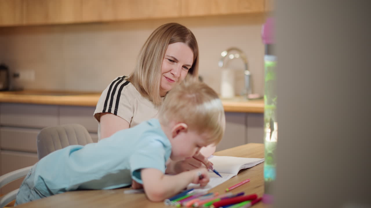 Mother watching young boy draw with colorful pens on notebook at wooden table beside bright aquarium, enjoying creative learning activity together in warm kitchen atmosphere with focus on education