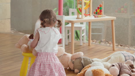 happy little child girl playing with her favorite teddy bear toy seating him down on stool at home