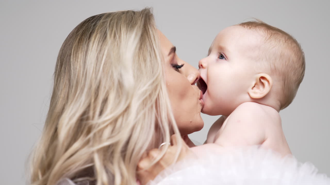 Loving mother with her beautiful infant baby. Blonde woman kissing her child and kid adorably opens his mouth. White backdrop.