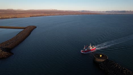 aerial drone view of red fishing boat moving through calm blue waters into harbor surrounded by rocky breakwaters, with open coastline, distant snowy mountains, and clear sky in iceland