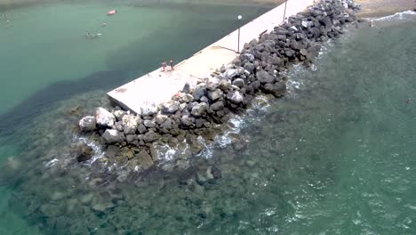 Aerial-view-of-stunning-turquoise-sea-in-the-summer-with-pier-and-rocks-that-water-pops-up-on-them-in-the-area-of-Agia-Paraskevi-Halkidiki,-Greece,-circular-movement-by-drone