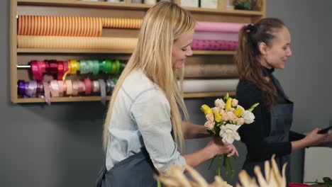 Smiling-blonde-florist-in-apron-standing-with-her-coworker-at-counter-in-floral-shot-while-arranging-bunch-of-flowers