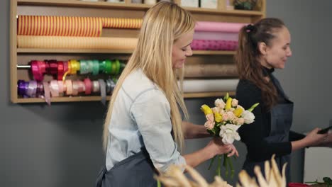 smiling blonde florist in apron standing with her coworker at counter in floral shot while arranging bunch of flowers