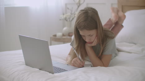 bored child girl is doing homework in copybook looking on laptop display during online lessons learning at home