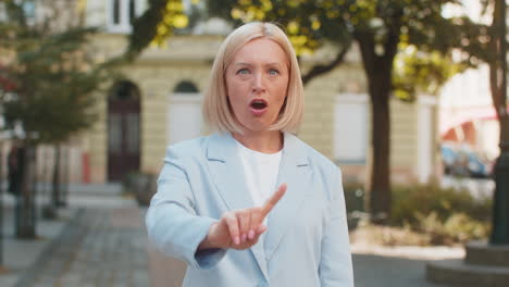 caucasian mature businesswoman in suit standing in city street looking at camera shaking finger