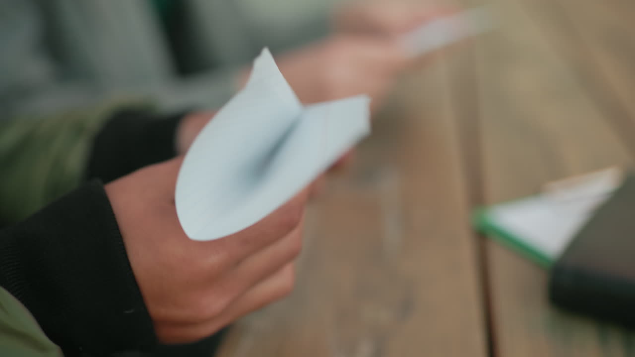 Close up of person folding paper into kite on wooden table with documents nearby, showing detail of hands engaged in craft activity with concentration and creativity in outdoor setting